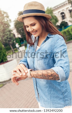 Young woman wearing hat walking outdoors in the city park looking at digital watch checking time smiling cheerful