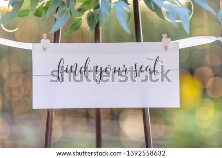 Find your seat sign for wedding or event white card with ribbon and wooden pegs and green leaf foliage above with blurred bokeh background and copyspace for names