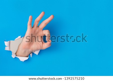 Male hand tearing through blue paper background creating ok gesture with hand and copy space for text. Royalty-Free Stock Photo #1392575210