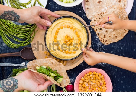 Adult woman and kid, child hands holds, eats creamy chickpea Israeli Arabic hummus dip paste with tahini, oil, garlic, paprika served with fresh vegetables, salad leaves and baked pita flat breads. Royalty-Free Stock Photo #1392601544