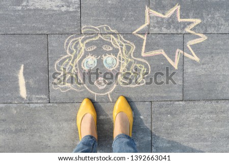 Abstract pop art woman face, picture written on gray sidewalk in crayons, with women legs in yellow shoes, top view
