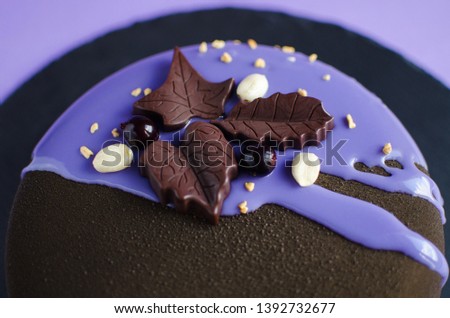 Contemporary mousse cake covered with brown velvet spray and violet mirror glaze, decorated with chocolate leaves, fresh black currant and peanuts on violet background