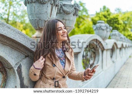 Beautiful woman texting on a smart phone in a city with a green background. Cheerful girl using smartphone in town. Smiling woman using mobile phone on the street 
