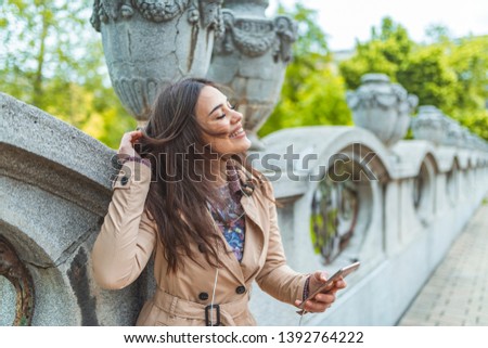 Young happy smiling girl using modern smartphone outdoors, beautiful woman texting sms message via her cellphone while walking at the park in the city. Woman  texting on the smart phone