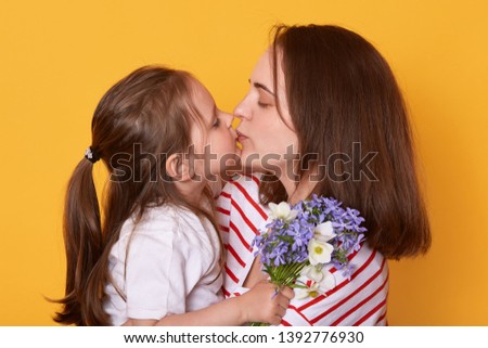 Studio shot of mother and daughter kissing and hugging, little girl standing with bouquet of blue florets for mommy. Dark haired woman poses with her child. Happy Mother's Day! Holiday concept.