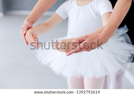 Ballet teacher helps little ballerina in classical dance school. Female dance trainer is teaching kid girl to do choreographic position with hands. Practicing for children. Close up. Royalty-Free Stock Photo #1392792614