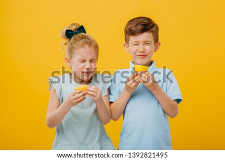 two young children, little girl and little boy taste sour lemon emotional, with sore, facial emotions negative, in blue T-shirt, isolated yellow background, copy space Royalty-Free Stock Photo #1392821495
