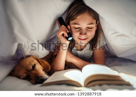 Girl lying in the bed with her small brown dog under blanket holding flashlight and reading book late at night Royalty-Free Stock Photo #1392887918