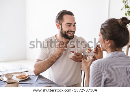 Image of joyous brunette couple man and woman 20s eating panna cotta dessert together while sitting at table at home