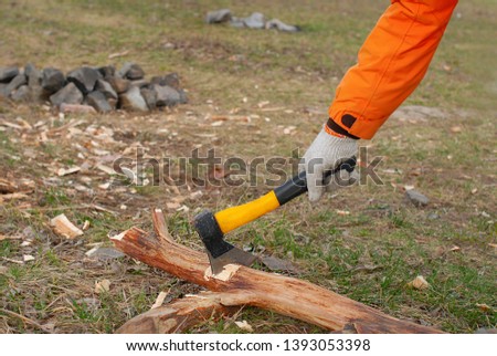 The man’s hand in a cloth glove and orange jacket holds an ax with which he cuts a dry log for firewood. The background is a stone fireplace, a picnic area.