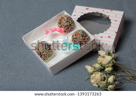Cake Pops in chocolate with colored sprinkles. Decorated with a ribbon bow. They lie in a gift box, in the lid of which there is a transparent window. Nearby are dried roses. On a gray background.