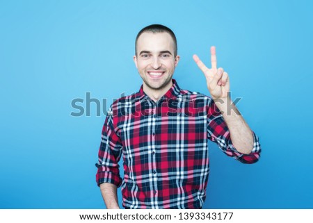 Attractive guy in plaid shirt smiling looking at camera, shows sign peace, front view, blue background, with copy space, for advertising, for slogan