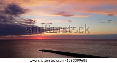 Scenic bright background. Sea evening sunset landscape. Clouds backlit by the setting sun, painted in red, pink, blue, purple tones. On a stone mall dark silhouettes of people 