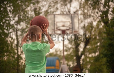 One nine years old boy, rear view from behind, holding a ball in his hands, raising it up to shoot. On a Spring day, at a basketball court outdoors.