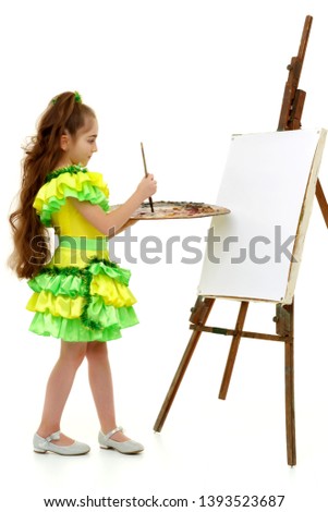 Cute little girl with a palette and brush near the easel. At the easel a blank white sheet of paper on which you can draw a picture or apply any advertising inscription. The concept of art, children's