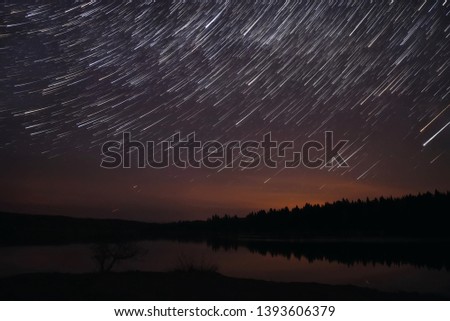 Amazing view of the night sky with stars in the form of tracks over the lake, forest and reflections in the water