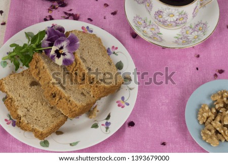 Three delicious slices of apple and walnut bread can be seen on a plate with edible flowers. A cup of black tea and a smaller blue plate of walnuts peep into the scene.  Rose petals are scattered.