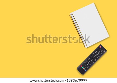 Black plastic remote control for tv near blank paper notepad with spiral binder lies on yellow table in home or office. Top view.Concept of communication or multimedia. Copy space for your text