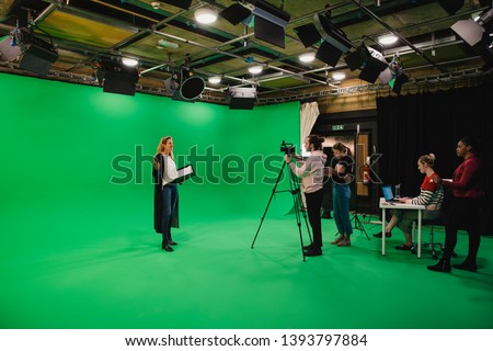 A wide-angle shot of a multi-ethnic group of people working in a film studio, a mature caucasian woman can be seen presenting in front of a green screen. 