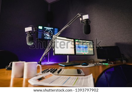 A front view shot of a radio station studio interior, a large desk is in the middle of the room, recording equipment and computers can be seen on the desk.