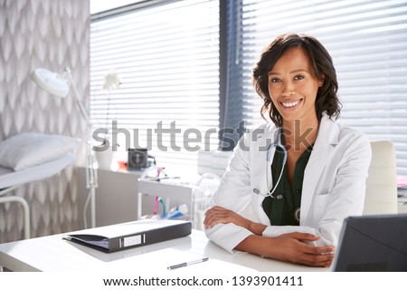 Portrait Of Smiling Female Doctor Wearing White Coat With Stethoscope Sitting Behind Desk In Office Royalty-Free Stock Photo #1393901411