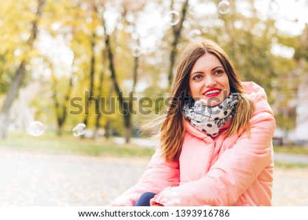Close up portrait of emorional woman in stylish pink down jacket and scarf. Autumn bright pink coat sitting in city sidewalk on street and enjoying amazing weekend time.