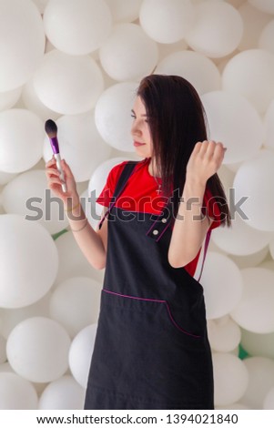 Professional make-up artist, young attractive brunette girl with long hair in uniform holding a brush in her hands, posing in a photo studio on a white background to advertise new cosmetics store