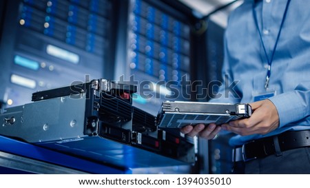 In the Modern Data Center: IT Engineer is Holding New HDD Hard Drive Prepared for Installing Hardware Equipment into Server Rack. IT Specialist Doing Maintenance and Updating Hardware. Royalty-Free Stock Photo #1394035010