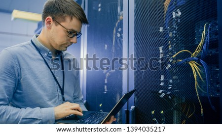 In Data Center IT Engineer Stands Before Working Server Rack Doing Routine Maintenance Check and Diagnostics Using Laptop. Visible Computer Hardware Equipment, Broadband Fiber Optic Cables LED Lights. Royalty-Free Stock Photo #1394035217