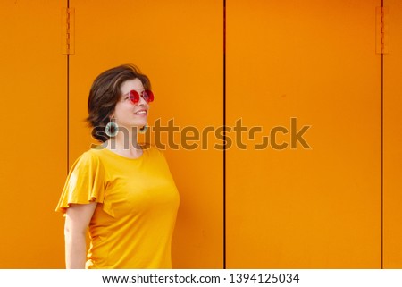 Beautiful young smiling woman wearing red sunglasses and yellow blouse standing on orange metal door. Happy girl having fun enjoying sunny day. Outdoor activities. Holiday or weekend spending time. 