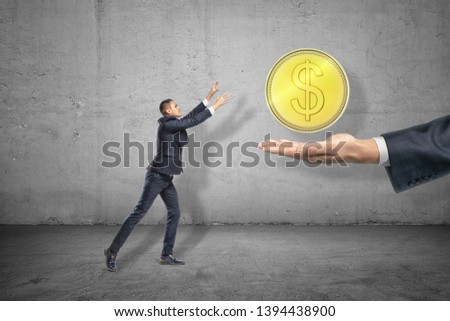 Side full length view of little businessman reaching out for big gold coin levitated above huge hand emerging from right. Greed for gain. Earning money. Gaining profits.