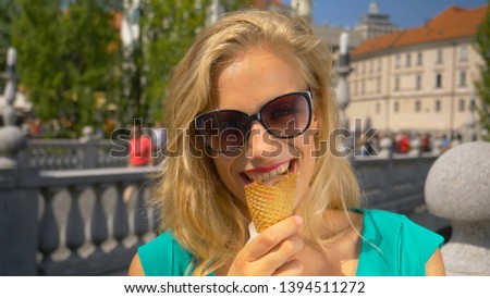 CLOSE UP, DOF, PORTRAIT: Happy Caucasian woman looks at the camera while eating delicious chocolate ice cream in the scenic streets of an old European town. Tourist girl eating an ice cream cone.