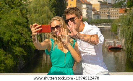 CLOSE UP, DOF: Happy travelers wave to the smartphone while shooting a travel vlog in beautiful sunny Ljubljana. Smiling girl and her boyfriend waving goodbye to their friends after a conference call.