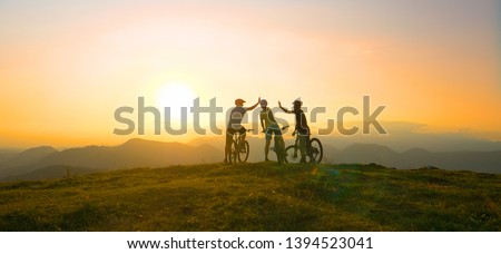 SUN FLARE: Mountain biking friends high five after reaching the scenic summit at breathtaking golden sunrise. Cross country cyclists celebrate a successful mountain biking adventure on a sunny evening Royalty-Free Stock Photo #1394523041