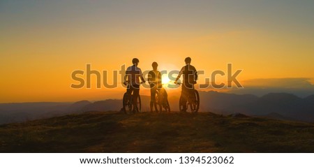 LOW ANGLE, SILHOUETTE, SUN FLARE: Golden evening sun rays illuminate cross country bikers on the mountaintop observing the scenic landscape. Three friends watch sunset after bicycle ride in mountains Royalty-Free Stock Photo #1394523062