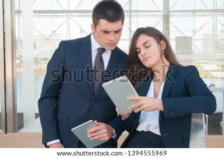 Pretty smiling woman in white blouse and serious young man in dark suite standing in office corridor, woman showing data on tablet. Work, communication concept