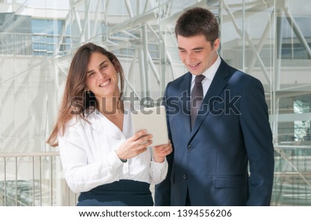 Pretty smiling woman in white blouse and serious young man in dark suite standing in office corridor, woman showing data on tablet. Work, communication concept