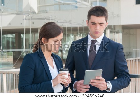Pretty woman in white blouse and serious young man in dark suite standing in office corridor, man showing data on tablet, woman looking, holding takeaway coffee in hand. Work, communication concept
