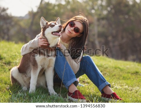 Young white girl and brown husky puppy chilling outdoor in green park.Royalty free photo of beautiful Caucasian brunette female model with long dark hair sitting at sunset with Siberian dog pup