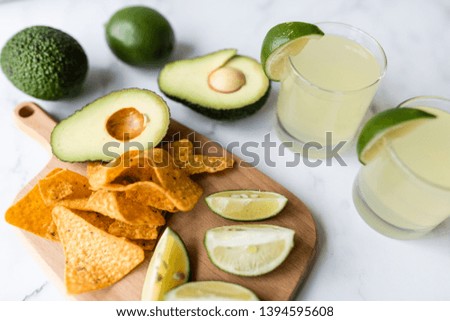 Fresh avocado, lime, drink and nacho chips lying on marble background. Recipe for Cinco de Mayo party