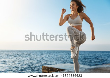 Motivated happy sporty woman wearing sports bra, sneakers enjoying excercise, training outdoors near sea, workout quay, jogging, running, jumping energized, smiling during productive fitness session Royalty-Free Stock Photo #1394684423