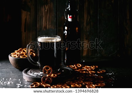 Dark craft beer in glass mug with empty beer bottle served with pretzels and salt over dark wooden background
