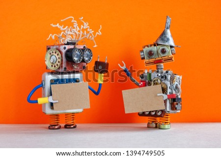 Two comical robots with a cardboards mockup. Creative design robotic toys holding a blank empty paper posters, orange wall background. copy space for text and design elements