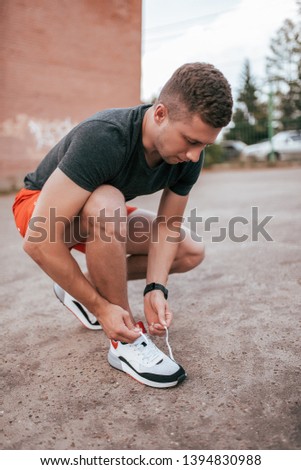Male athlete runner tying shoelaces on sneakers in the city in the fresh air. Fitness concept and workout, smart watch bracelet. Sportswear T-shirt shorts.