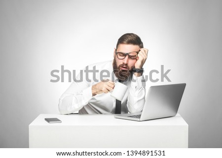 Irresponsible tired young manager in white shirt and black tie is sitting in office and trying not to sleep on the work, drink a cup of coffee, holding head with hand. Studio shot, isolated, indoor Royalty-Free Stock Photo #1394891531