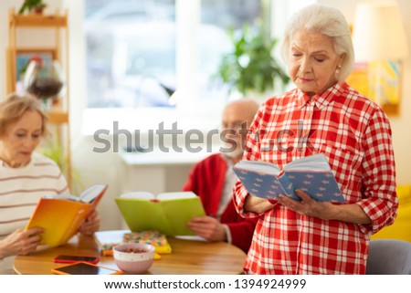 I like reading. Serious grey haired woman standing with a book while reading it