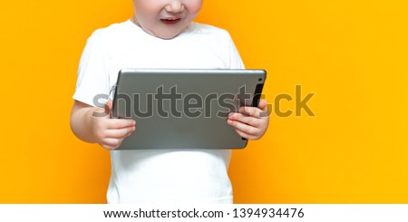 Surprised blonde three years old boy with his mouth open surprised, holding in his hands a tablet pc and looking at the camera on yellow background.