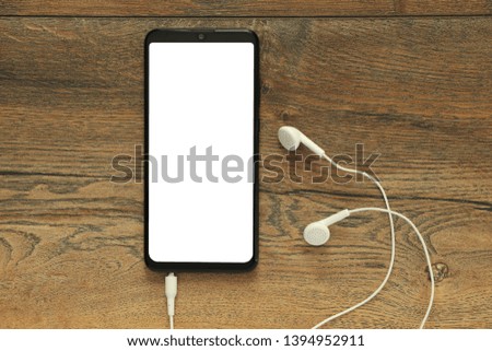 Smartphone with isolated white blank screen with connected earphones on the background of a wooden desk