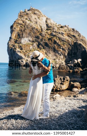 the guy kisses a girl in a white dress and a hat on the beach against the backdrop of the rocks