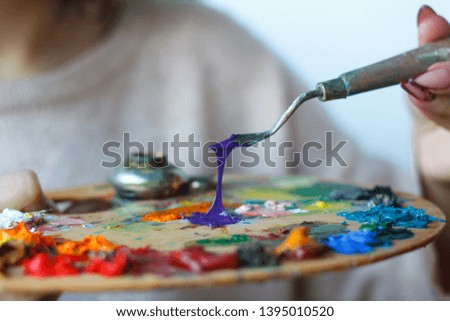 Close-up of female hands mixing paints on a palette with a spatula against a white canvas, creating an oil painting, soft focus
